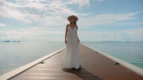 Young Lovely Woman in Long White Dress and Hat Walking on the Wooden Pier on Sunny Calm Morning