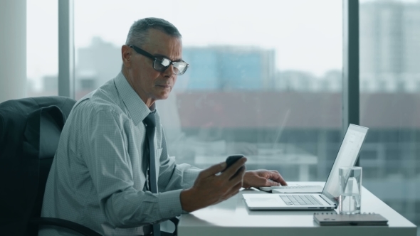 Elderly Businessman Look At Phone And Working With Computer In Modern Office