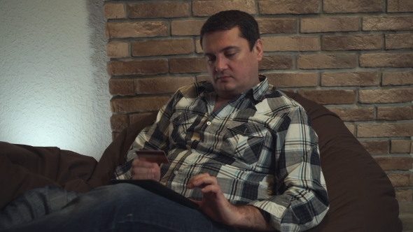 A Man Sitting In a Chair Makes a Purchase On His Tablet. alt