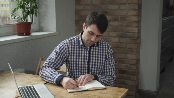Attractive Man In a Shirt Making Notes In a Diary And Looking At a Laptop. alt