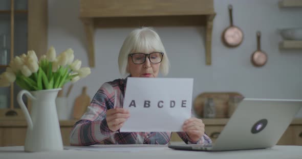 Senior Female Teacher in Glasses Talking and Showing Sheet of Paper with Alphabet Letters Looking