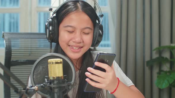 Asian Kid Girl Holding Phone And Wearing Headphones Singing While Recording Podcast In Studio