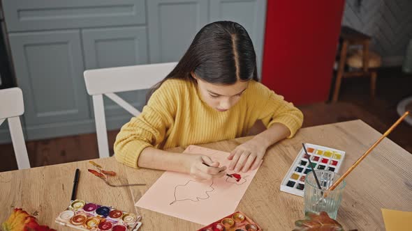 Preteen Girl Drawing Autumn Leaves with a Brush and Orange Watercolor Paint While Sitting at Table