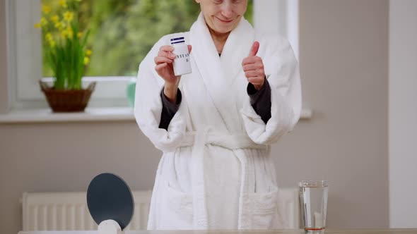 Unrecognizable Slim Mature Woman in White Bathrobe Gesturing Thumb Up Smiling Showing Bottle with