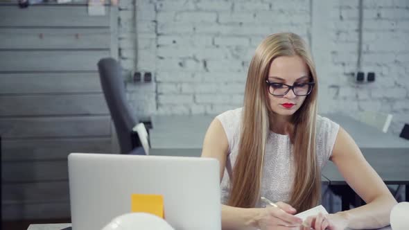 Beautiful Businesswoman Writing on Document Sitting at Table with Laptop in Office Interior.