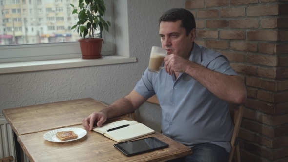 Adult Male In The Morning At The Hotel Drinking Coffee And Eating Toast, Is On The Table And Tablet alt