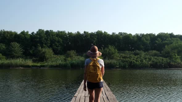 Young Tourist Woman in Hat and with a Backpack is on a Wooden Bridge Over the River