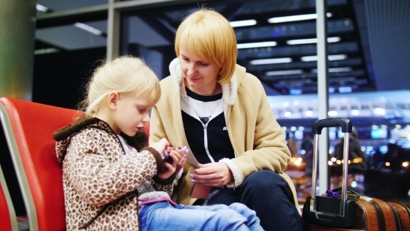 Girl 5-6 Years, Use Your Phone In The Airport Terminal, Next To Her Mother Sitting