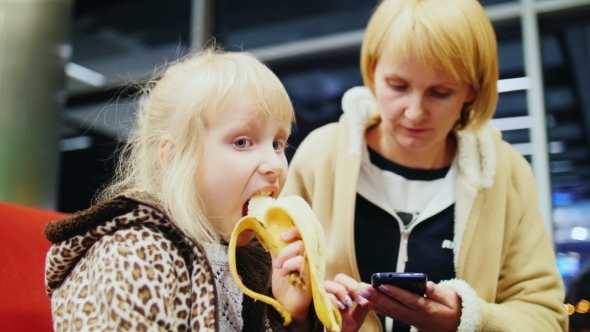 Little Girl Eating a Banana At The Airport With Her Mother Who Writes SMS alt