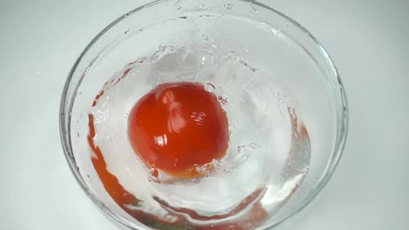 A ripe red tomato falls into a glass bowl and sprays water, slow motion