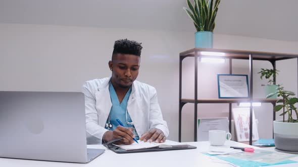 Black Man Writing Medical Notes and Drinking at the Desk Wearing Medical Gown