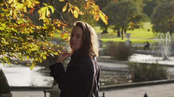 Caucasian woman with brunette long hair looking at camera under tree with shiny urban pond water and