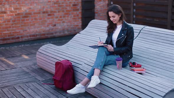 Female Student Writing in Notebook Sitting Outside on the Modern Wooden Bench