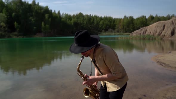 Male Musician Is Playing Saxophone Outdoor at Shore of Forest Lake at Summer Day
