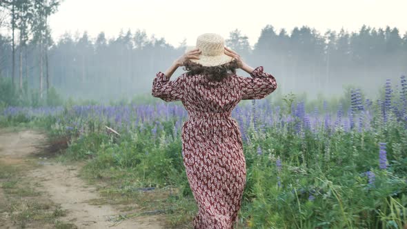 Girl in Dress Puts on Straw Hat and Runs Along Lupin Field