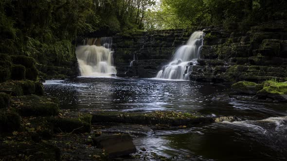 Time lapse of dark spring forest cascade waterfall surrounded by trees with rocks in the foreground alt