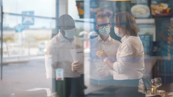 View Through Window of Multiethnic Waiters in Safety Mask and Gloves Wearing Uniform Discussing Menu