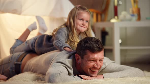 Portrait of Laughing Caucasian Father Lying at Tent in Living Room with Daughter on Back