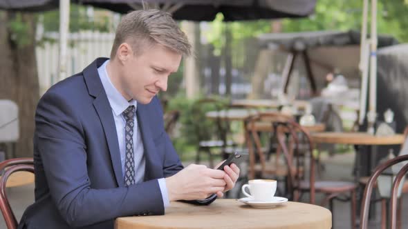Outdoor Sitting Businessman Celebrating Results on Smartphone