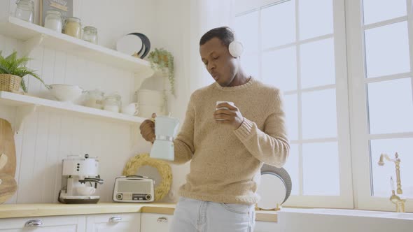 Cheerful Black Man Listening to Music and Dancing at Home in the Morning in the Kitchen and Drinking