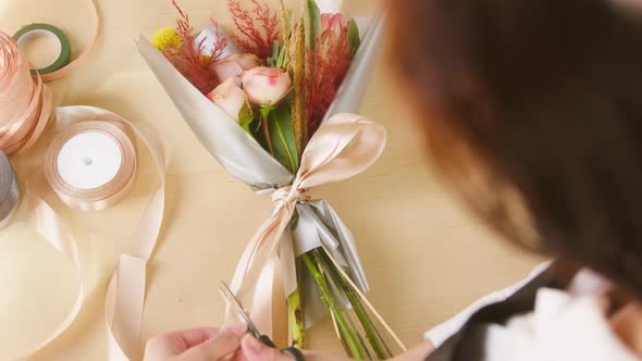 Young Florist Cutting Ribbon on Wrapped Bouquet of Roses and Dried Flowers with Packaging Paper at