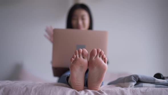 Feet of Asian Woman Sitting on Bed with Blurred Smiling Millennial Waving and Talking at Video Chat