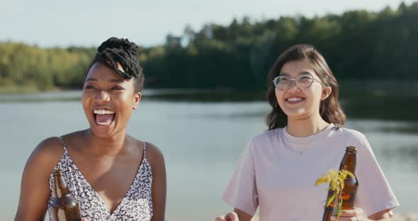 Two Beautiful Smiling Friends are Spending Time Together at the Lake Shore Holding Glass Bottles