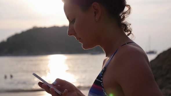 Woman Using Mobile Phone On Beach By Sea At Sunset