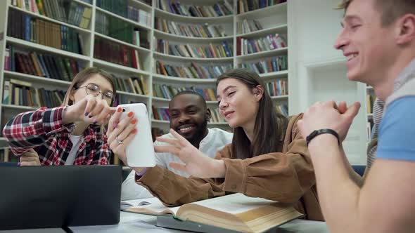 Young Multiracial People Sitting in Library and Smiling from Funny Videos on Girl's Phone