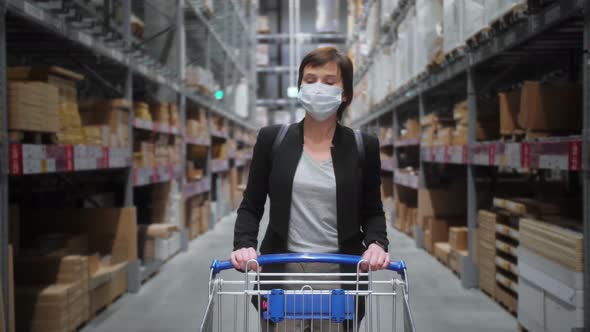 Woman in medical mask walking in mall steering shopping trolley looking around at shelves with goods