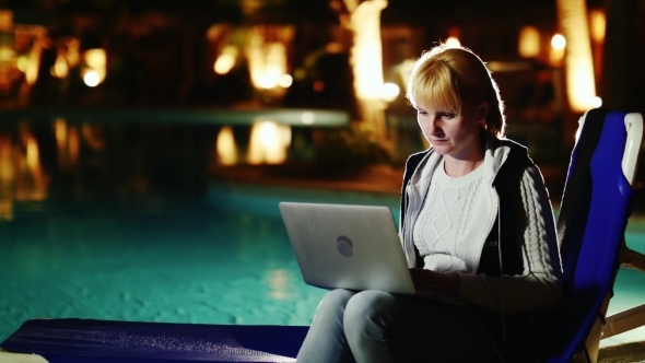 Woman Sitting Near The Pool In The Evening, Enjoying a Laptop
