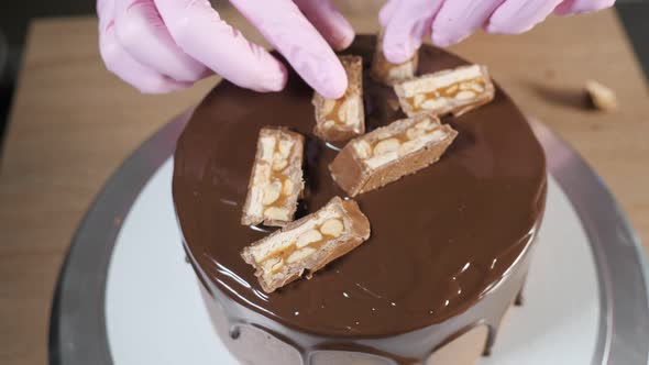 Closeup Pastry Chef's Hands is Decorating the Cake with Chocolate Pieces and Peanuts