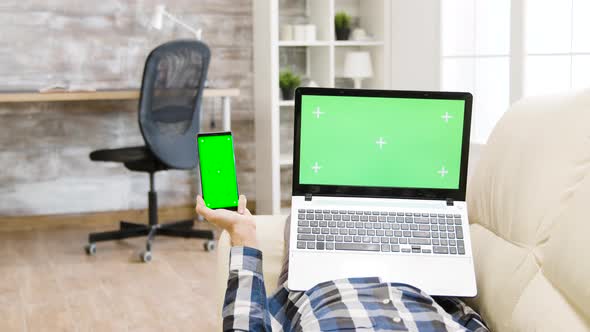 Parallax Shot of Man Lying on the Couch, Holding a Green Screen Smartphone and Laptop in Hands