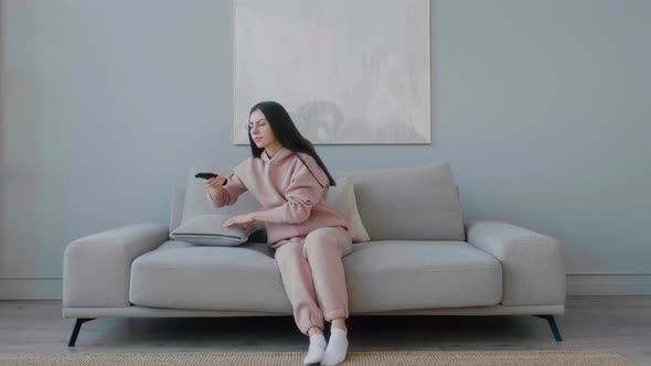 Young Caucasian woman with glasses wearily sitting down on sofa with a remote control in her hands.