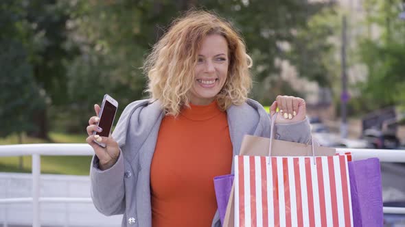 Positive Young Red-haired Woman with Shopping Bags and Cellphone in Hands Jumping and Rising Hands