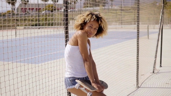 Young Woman With Skateboard Leans Against Fence