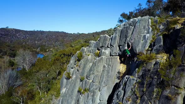 Stationary/hovering drone footage of an adult male rock climbing with a scenic creek and mountain in