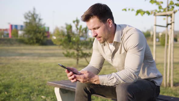 A Young Caucasian Man Holds a Smartphone and Talks to the Camera As He Sits in a Park
