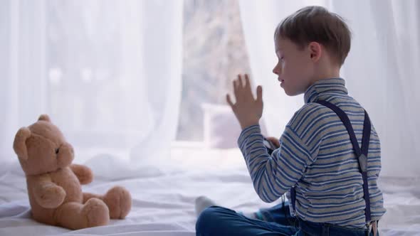 Curios Autistic Concentrated Boy Taking Selfie on Camera Sitting on Bed with Teddy Bear