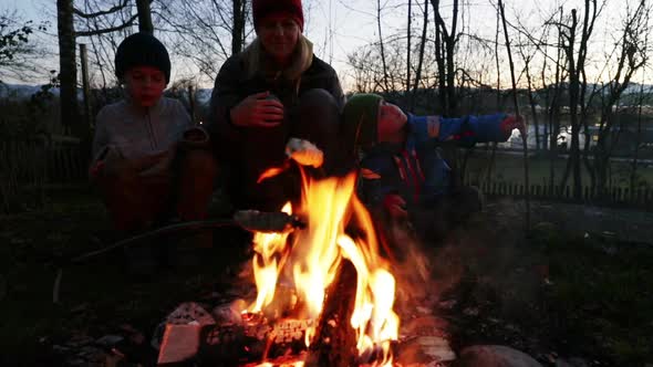 Two boys and their mother sitting around a bonfire with stick bread