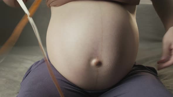 Young Pregnant Woman Sitting on Sofa Measuring Belly with Tape Measure Indoors