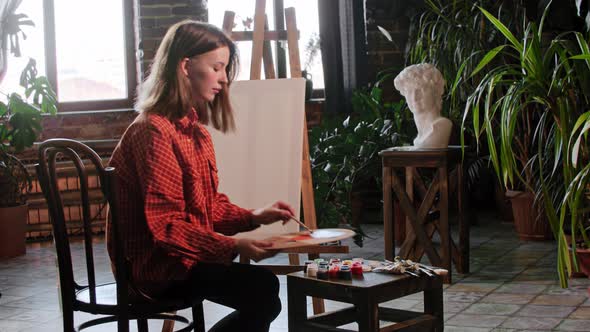 A Young Pretty Woman Artist Sits in Front of an Easel and Starts Mixing Colors on a Palette