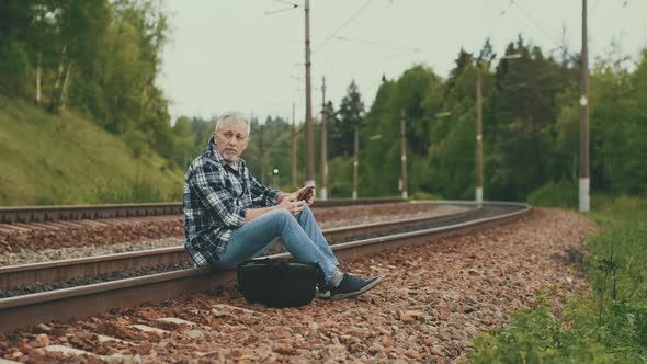 An Elderly Male Tourist Sitting on the Railway Tracks Looking at the Information on the Phone and