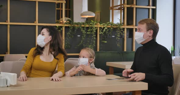 Young Caucasian Couple of Parents and Daughter in Covid19 Face Masks Sitting in Restaurant As Waiter