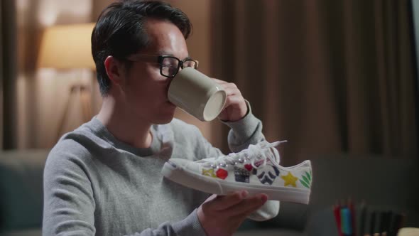 Asian Male Footwear Designer Drinking Coffee While Working On A Desktop Computer At Home