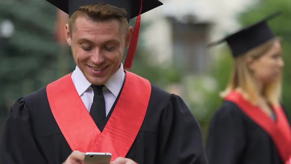 Cheerful Male Graduate in Academic Dress Chatting on Smartphone and Laughing