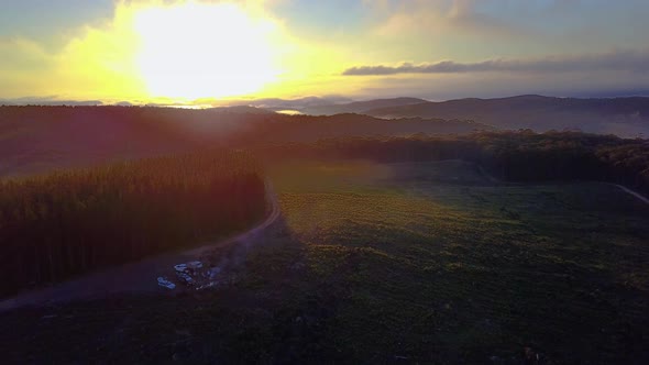 Drone footage of sunrise over a forest covered in patches of fog. People can be seen camping in bott