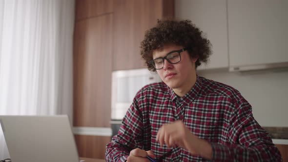 Curly Haired Male Student Attractive Young Boy in Glasses is Studying at Home Using Laptop Typing