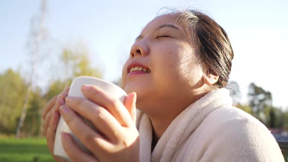 Asian woman holding and drinking hot coffee at a park near her house in the morning