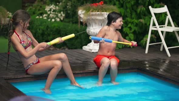 Wide Shot of Joyful Couple of Kids Having Fun Playing with Water Gun Sitting on Poolside Outdoors